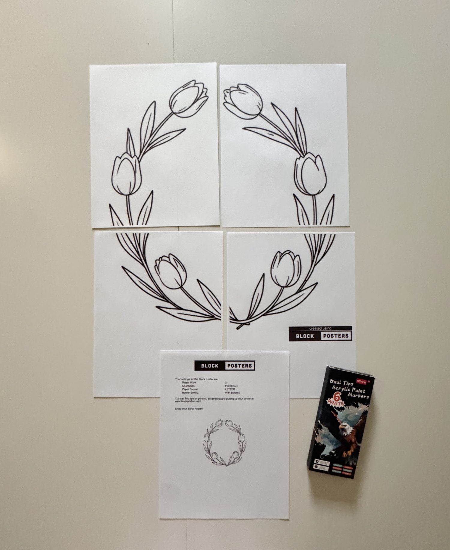 Four sheets of paper with parts of a tulip wreath outline are arranged on a table, along with an instruction sheet and a box of black poster markers.