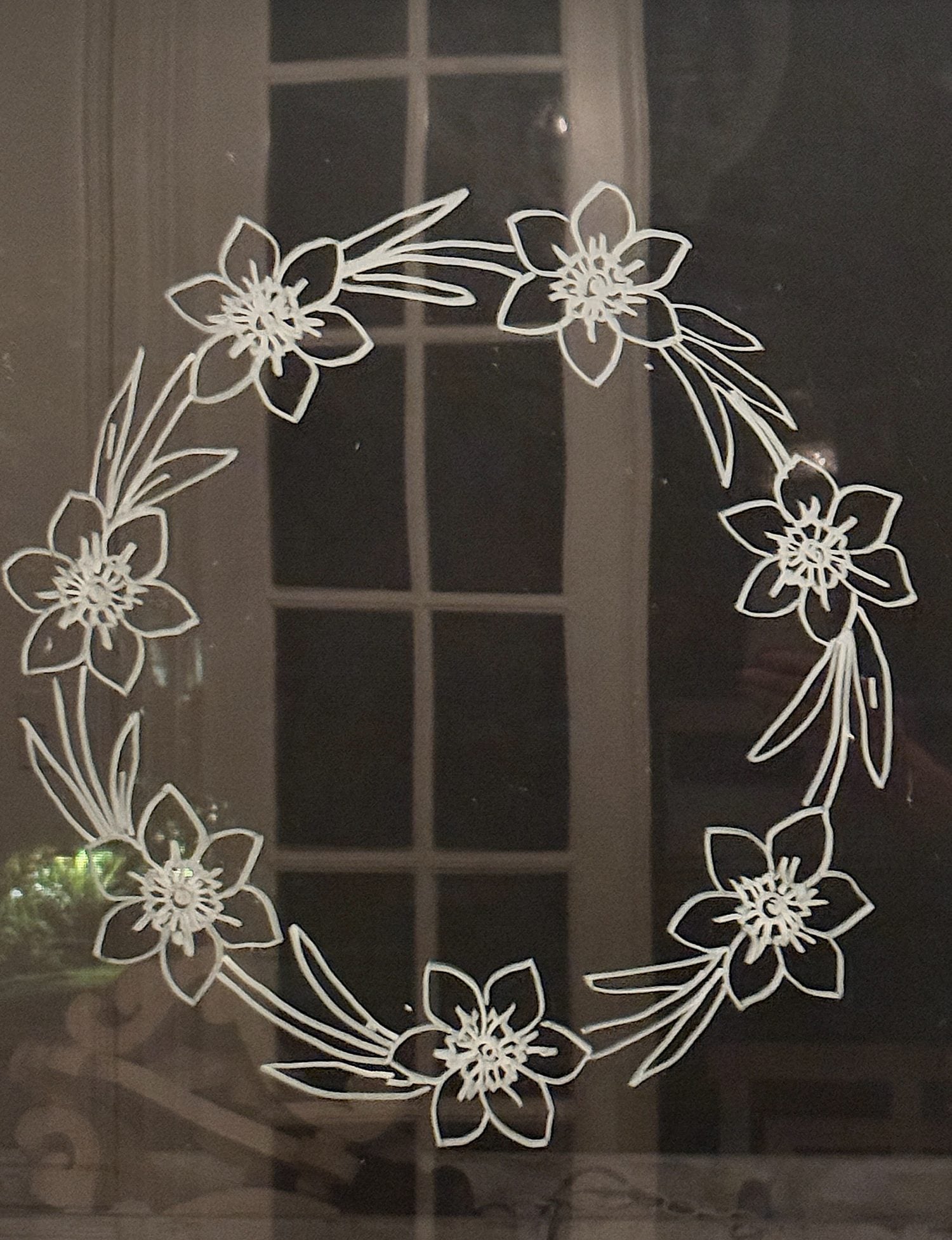 A white floral wreath design with six flowers and leaves is drawn on a glass surface with a window in the background.