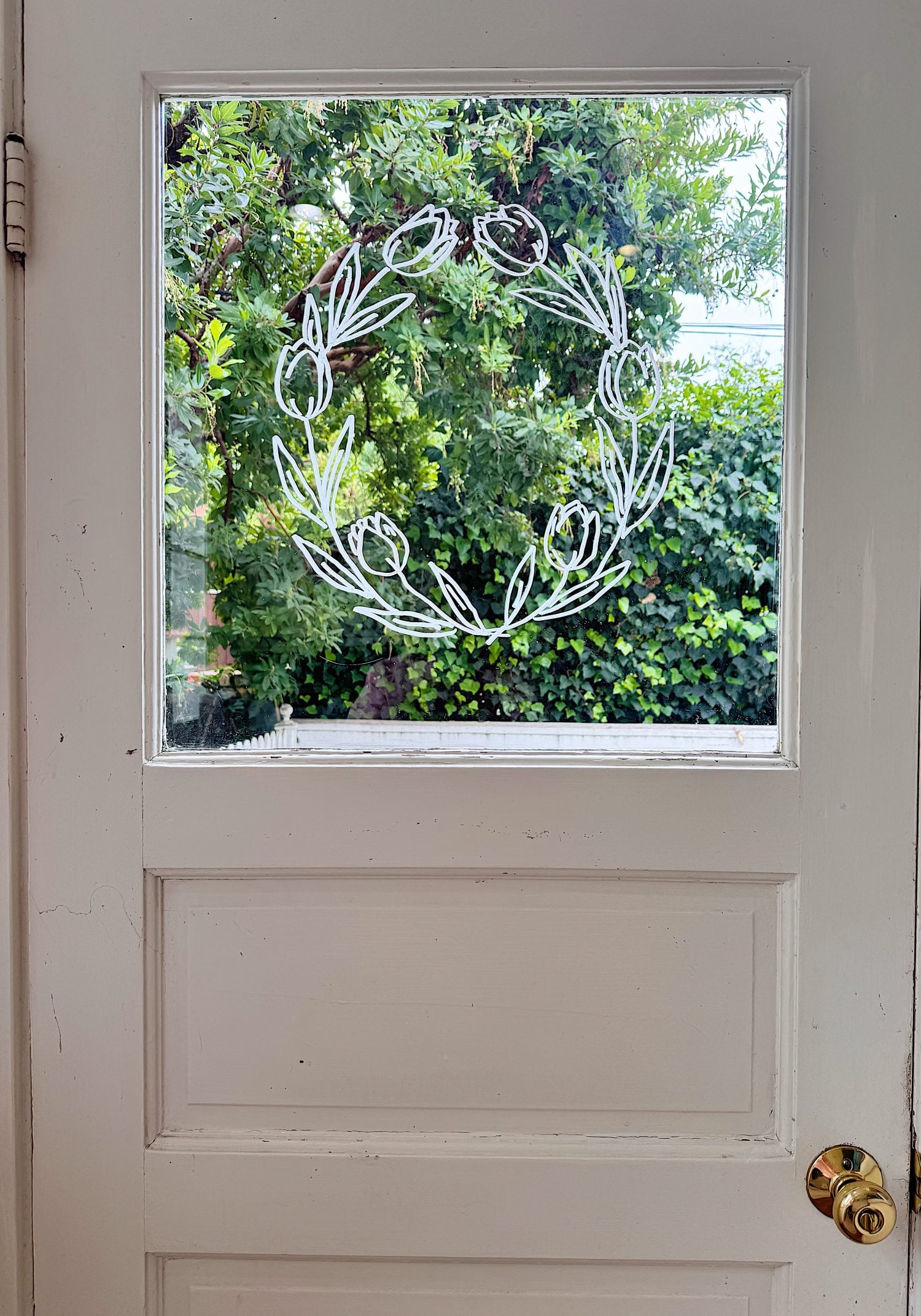 A white door with a glass window pane featuring a simple white marker drawing of a wreath; greenery is visible outside.