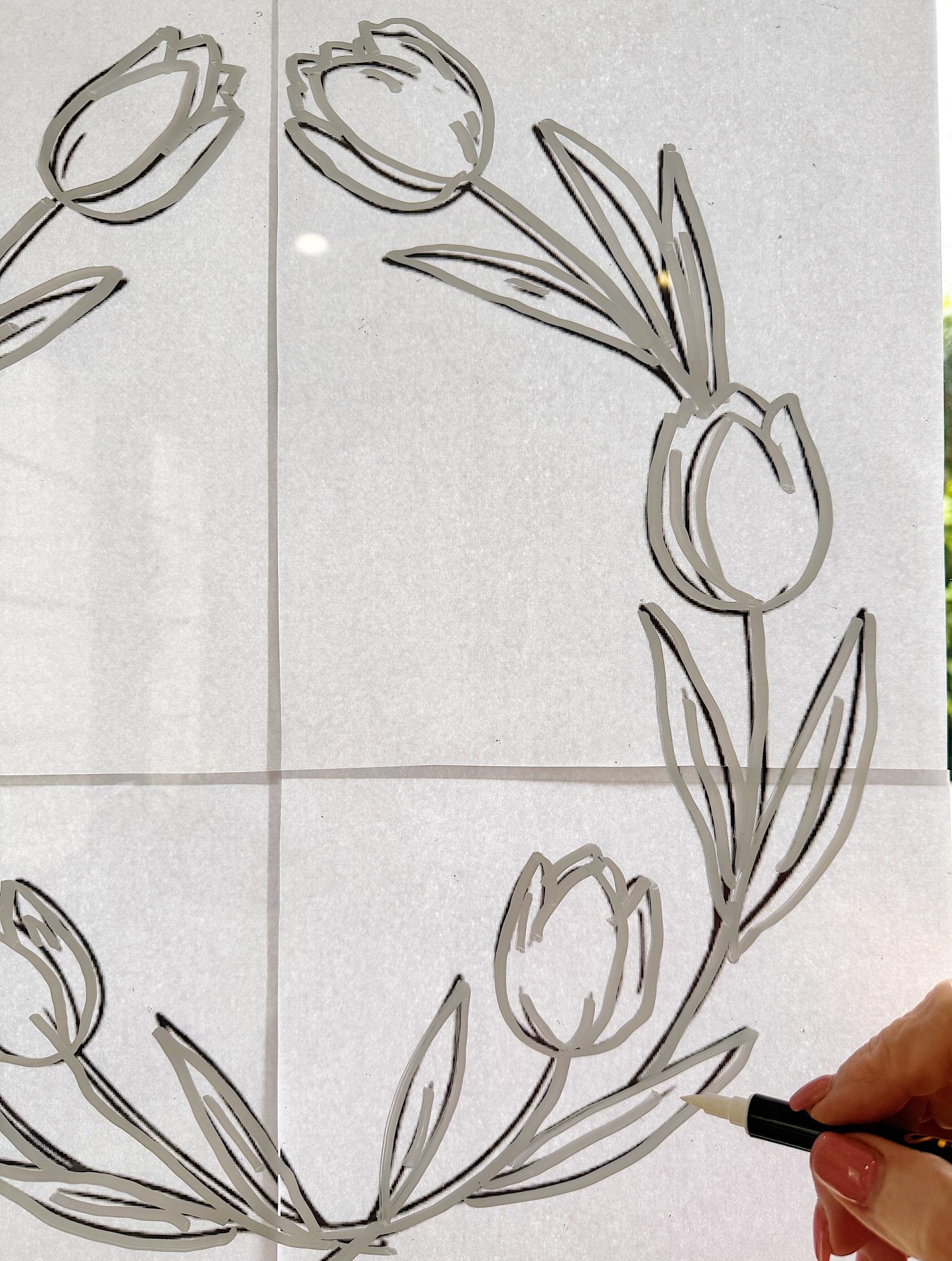 A hand uses a black marker to trace a tulip wreath design onto a window or glass surface from a paper template taped behind it.