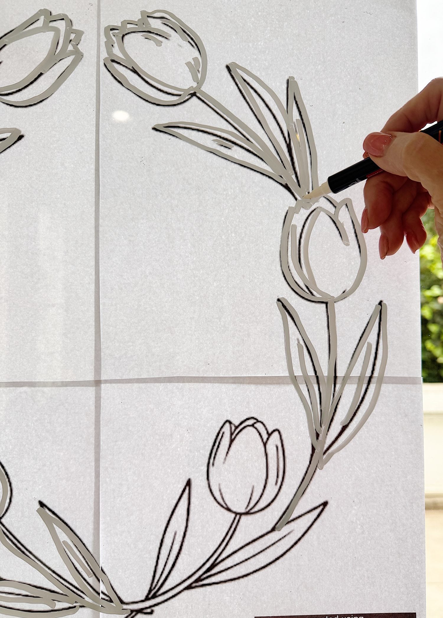 A hand traces tulip flowers and leaves with a black marker on a transparent surface, using a floral design template underneath.