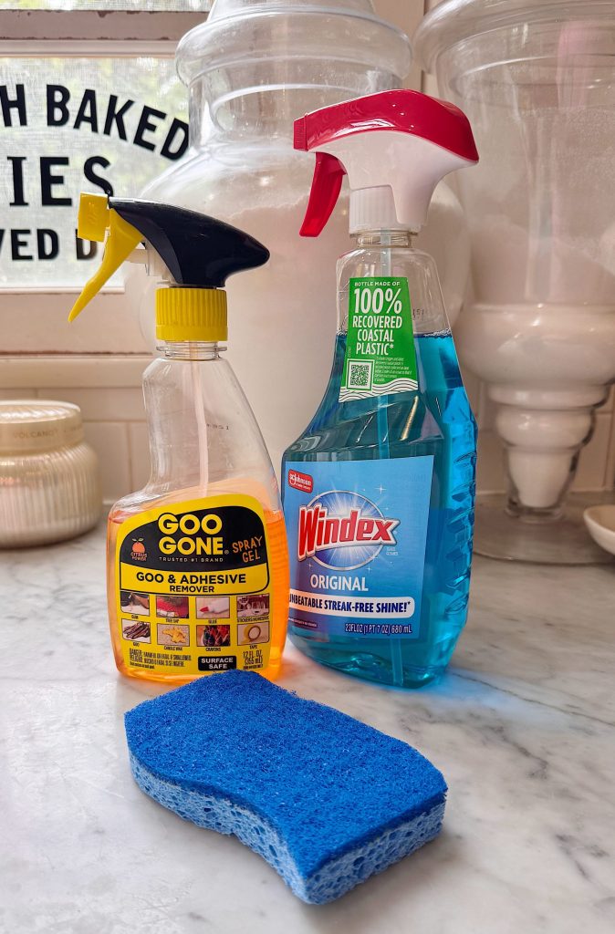 A bottle of Goo Gone, a bottle of Windex, and a blue sponge sit on a marble countertop with glass containers in the background.