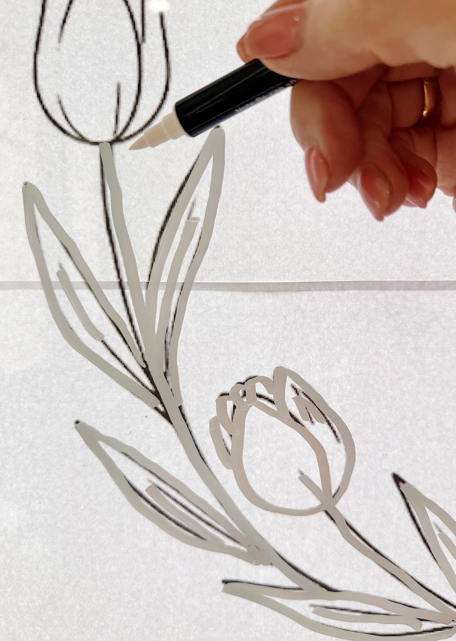 A hand with painted nails traces a black outline of tulips on a backlit surface using a marker.