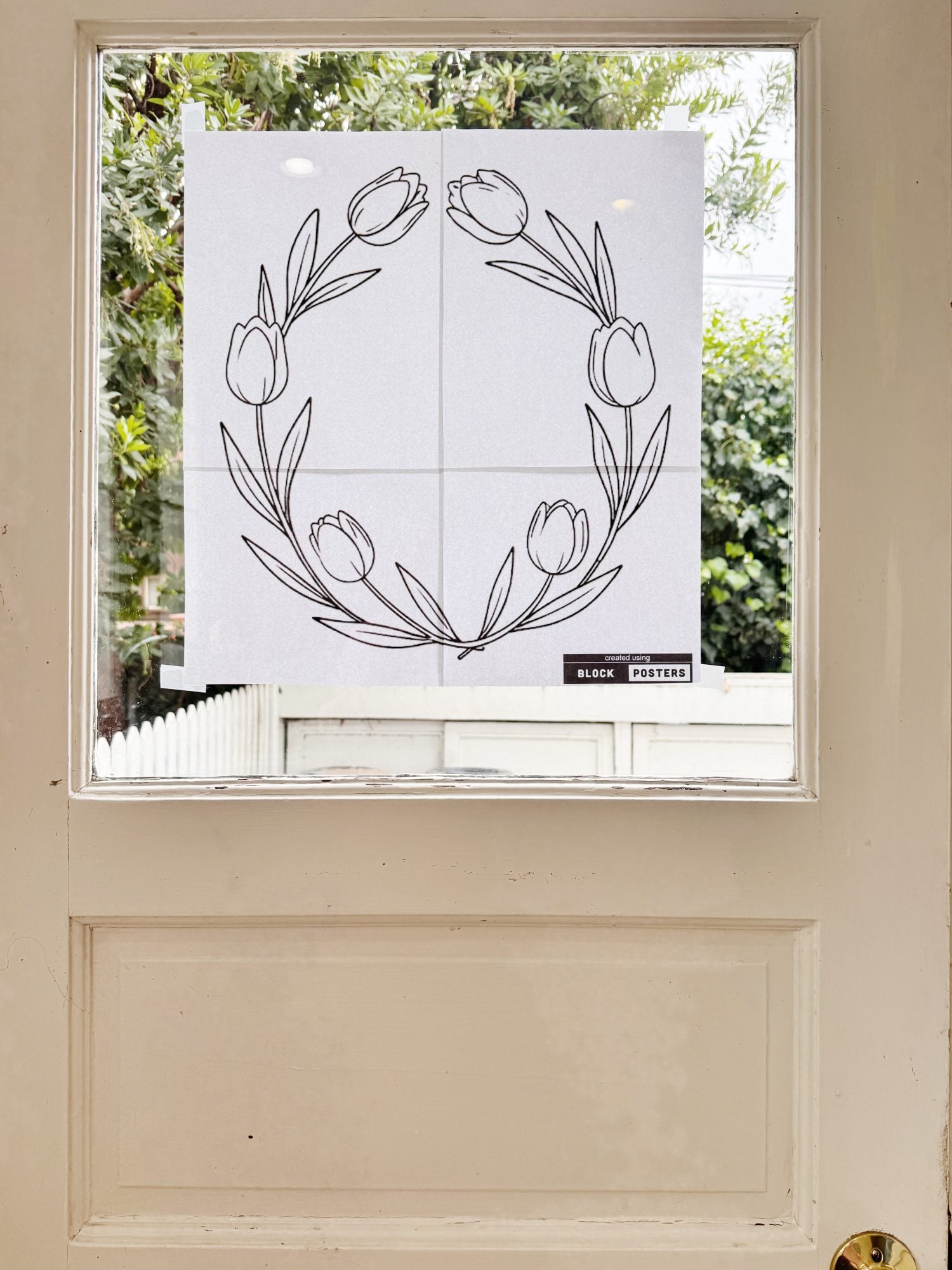 A black and white floral wreath outline is taped to a glass door, with trees and a white fence visible outside.