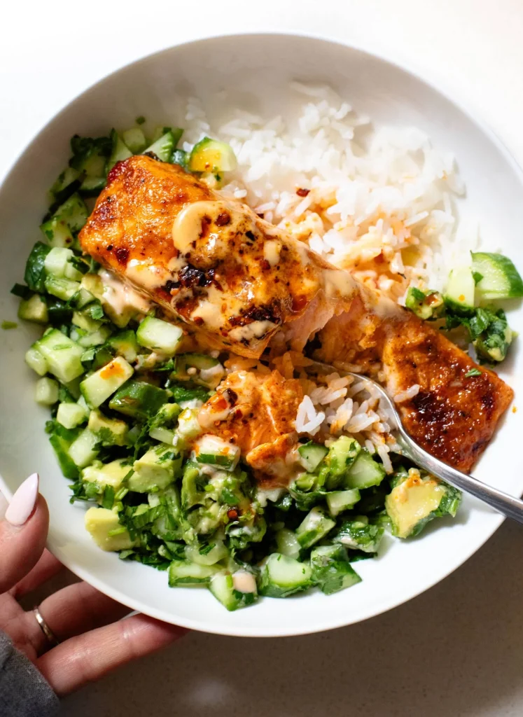 A bowl with baked salmon, white rice, and a cucumber avocado salad, with a fork and a hand holding the bowl.