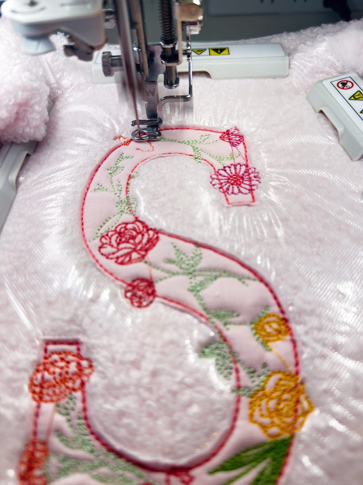 Close-up of a sewing machine embroidering a large, floral-patterned letter "S" on a piece of light pink, textured fabric.