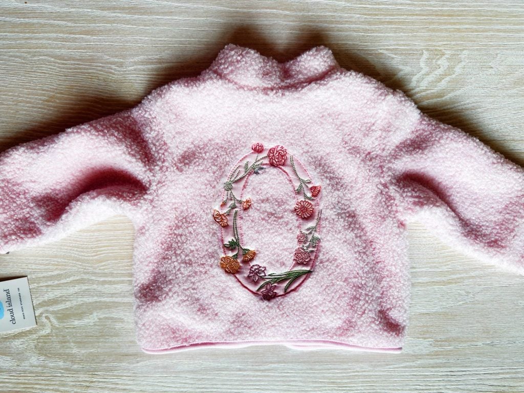 A pink, fuzzy baby sweater with long sleeves and floral embroidery forming an oval design on the back, displayed on a light wood surface.