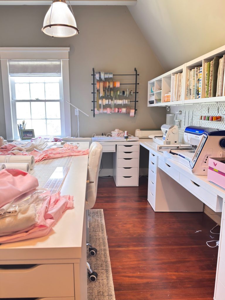 A sewing room with white furniture, fabric on the desk, sewing machines, shelving with fabric rolls, and ribbons hanging on the wall near a window.