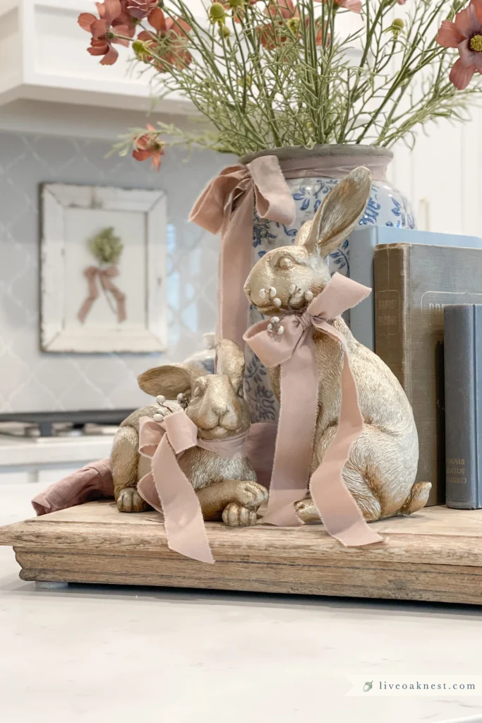 Two decorative gold rabbit figurines with pink ribbons sit on a wooden tray next to books and a blue floral vase filled with orange flowers on a kitchen counter.