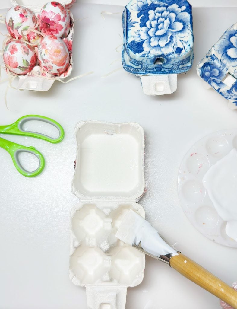 Egg cartons with floral-patterned eggs, a pair of green scissors, a paintbrush with white glue, and a paint palette on a white surface.