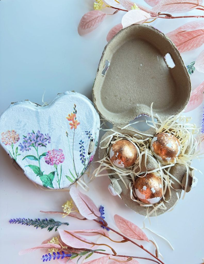 A heart-shaped egg carton decorated with floral designs holds straw and three metallic eggs, surrounded by pastel leaves and dried flowers.