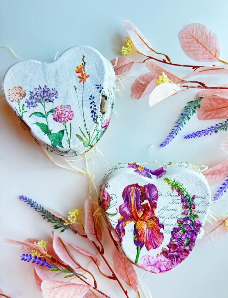 Two heart-shaped boxes decorated with floral designs are surrounded by pink leaves and sprigs of purple flowers on a white surface.