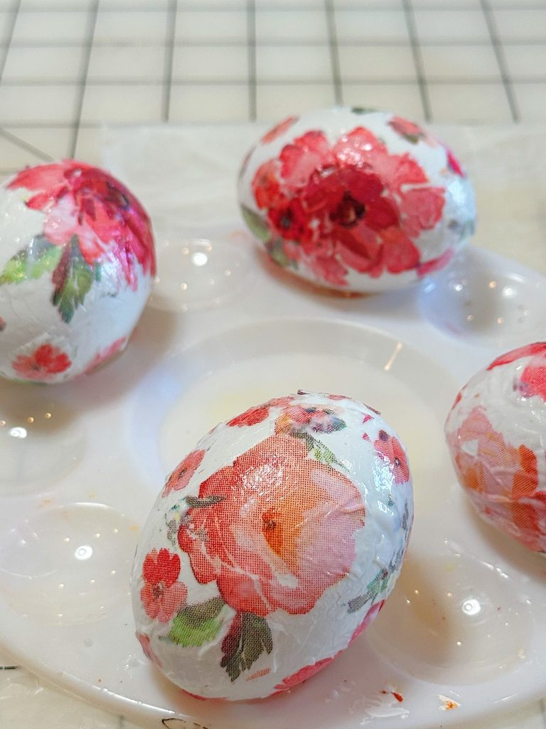 Four eggs decorated with white and red floral patterns rest on a plastic paint palette, with a tiled surface in the background.