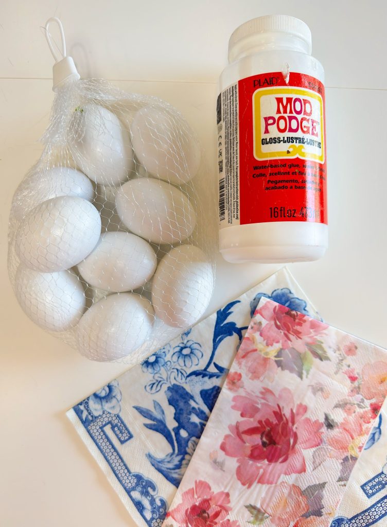 A bottle of Mod Podge glue, a mesh bag of white eggs, and two patterned napkins with floral and blue designs on a white surface.