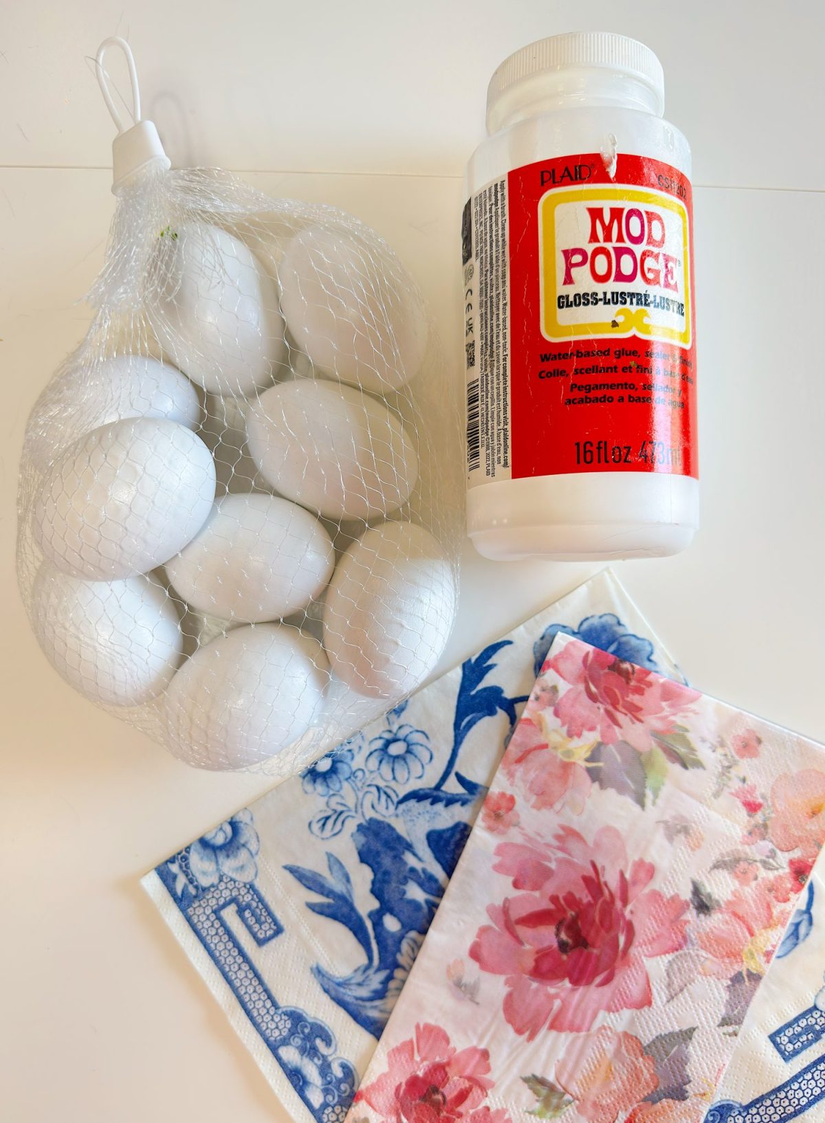 A bottle of Mod Podge glue, a mesh bag of white eggs, and two patterned napkins with floral and blue designs on a white surface.