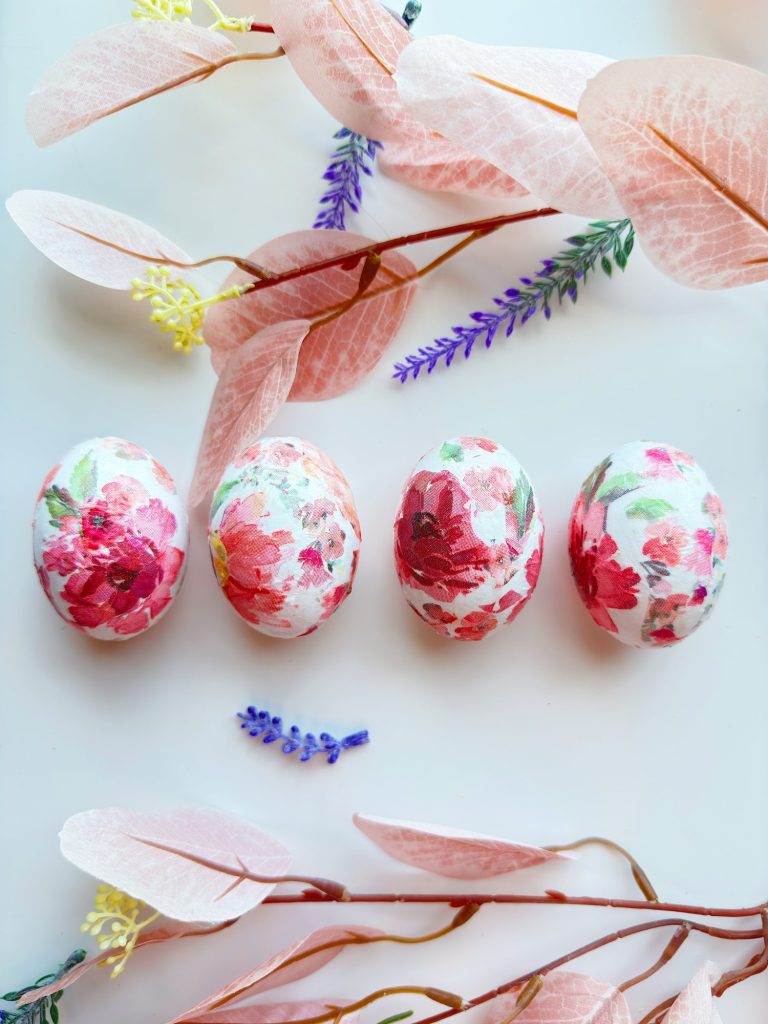 Four eggs decorated with red and pink floral patterns are arranged in a row on a white surface, surrounded by pink leaves and small purple flowers.