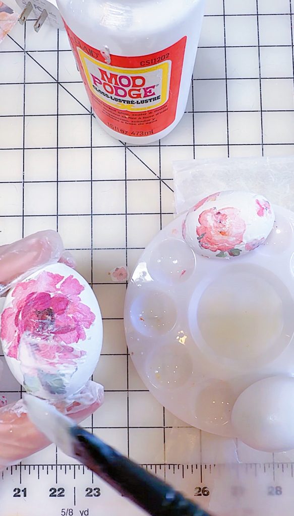 A gloved hand applies decoupage glue to a white egg with a pink floral design next to a paint palette, another decorated egg, and a bottle of Mod Podge on a grid surface.