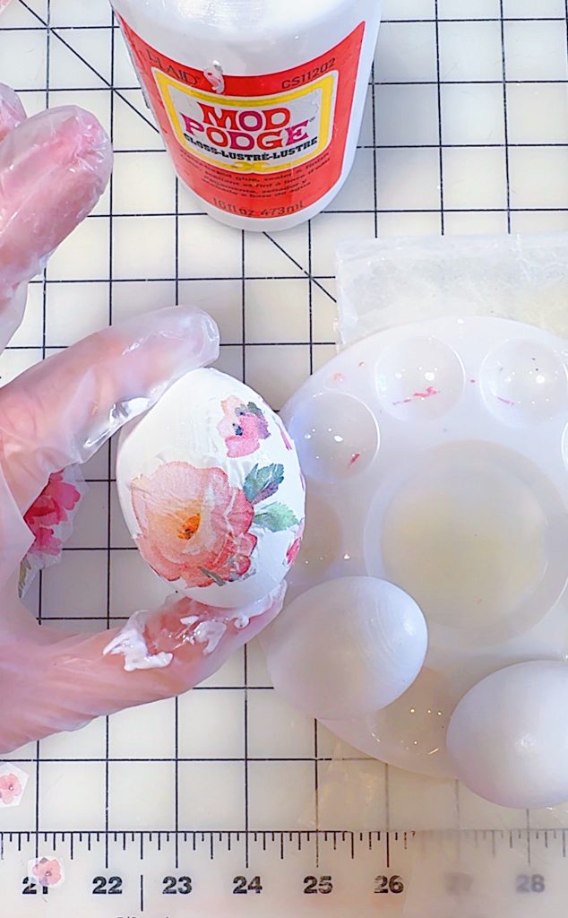 A gloved hand applies floral decoupage to an egg using Mod Podge, with a palette, two plain eggs, and a ruler on a grid-patterned workspace.
