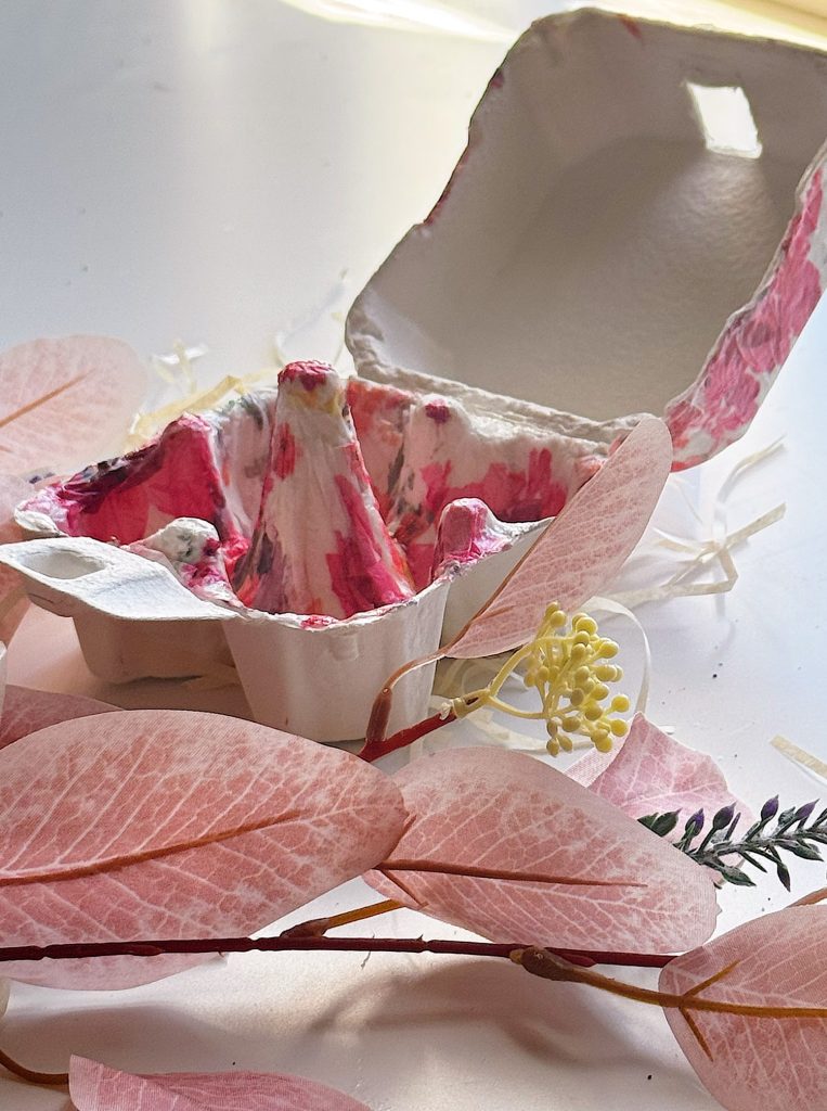 An open, empty egg carton with pink paint stains sits on a white surface, surrounded by artificial pink leaves and a yellow flower.