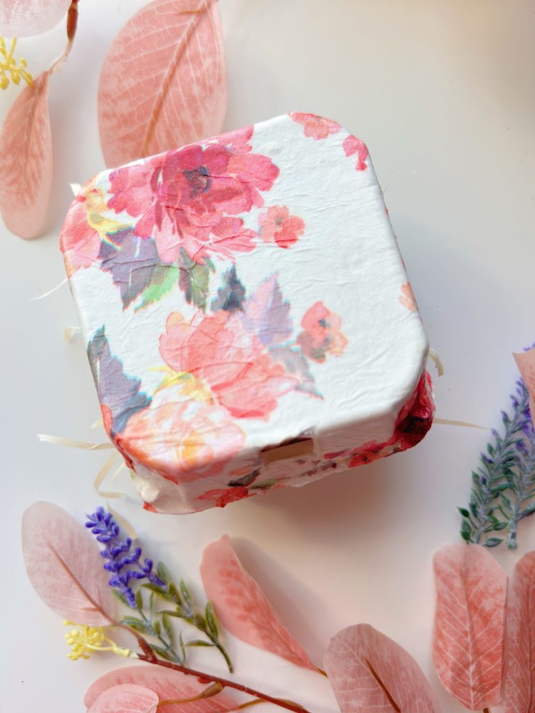 A small square box with a floral pattern sits on a white surface, surrounded by pink leaves and sprigs of lavender.