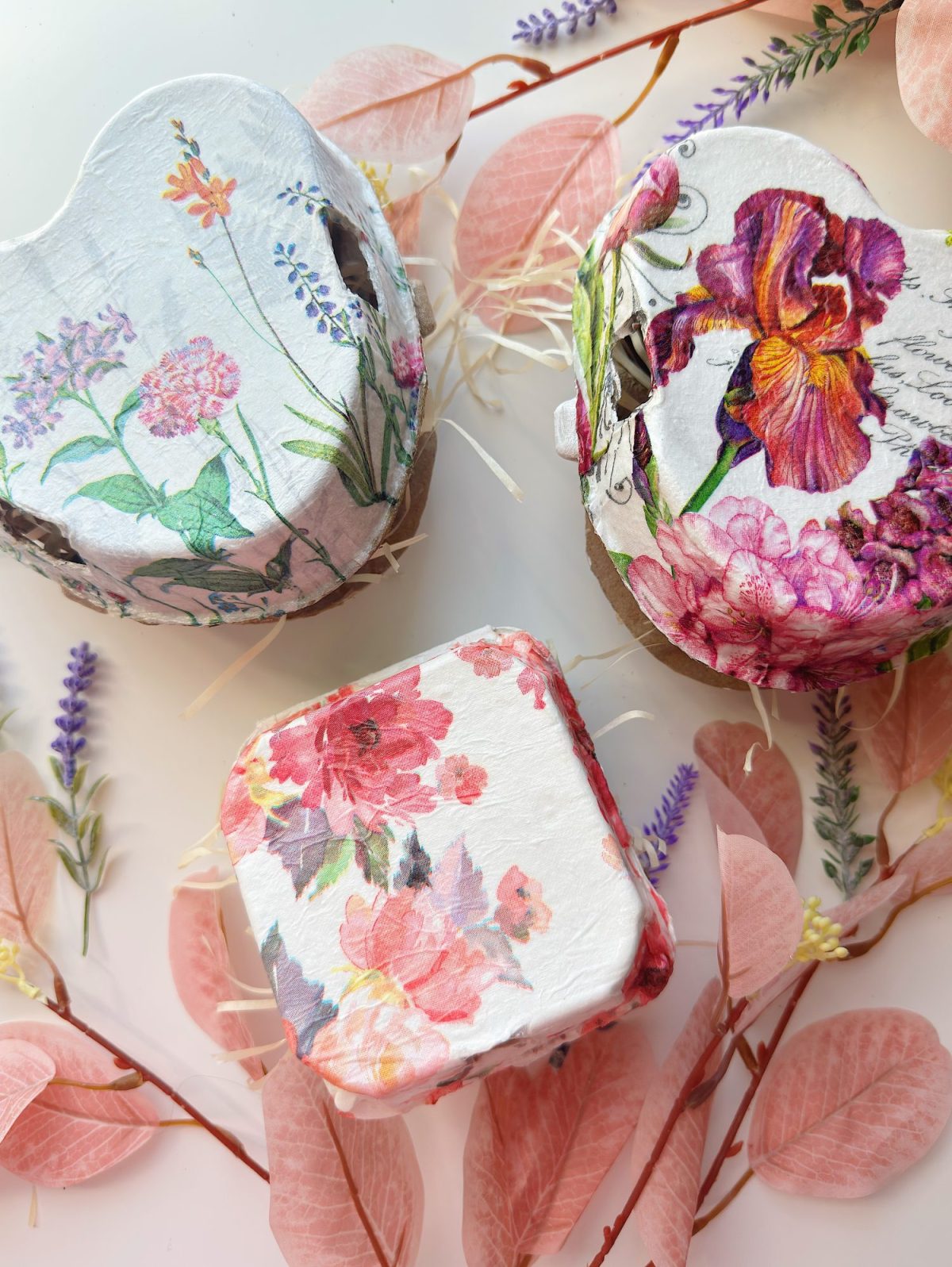 Three decorative boxes with floral designs are arranged on a white surface, surrounded by pink and purple artificial leaves and flowers.