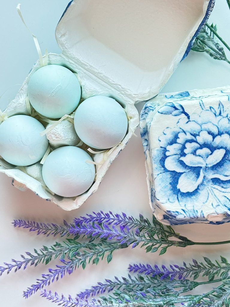 Four pale blue eggs in an open carton next to a closed carton with a blue floral design, with sprigs of lavender placed nearby.