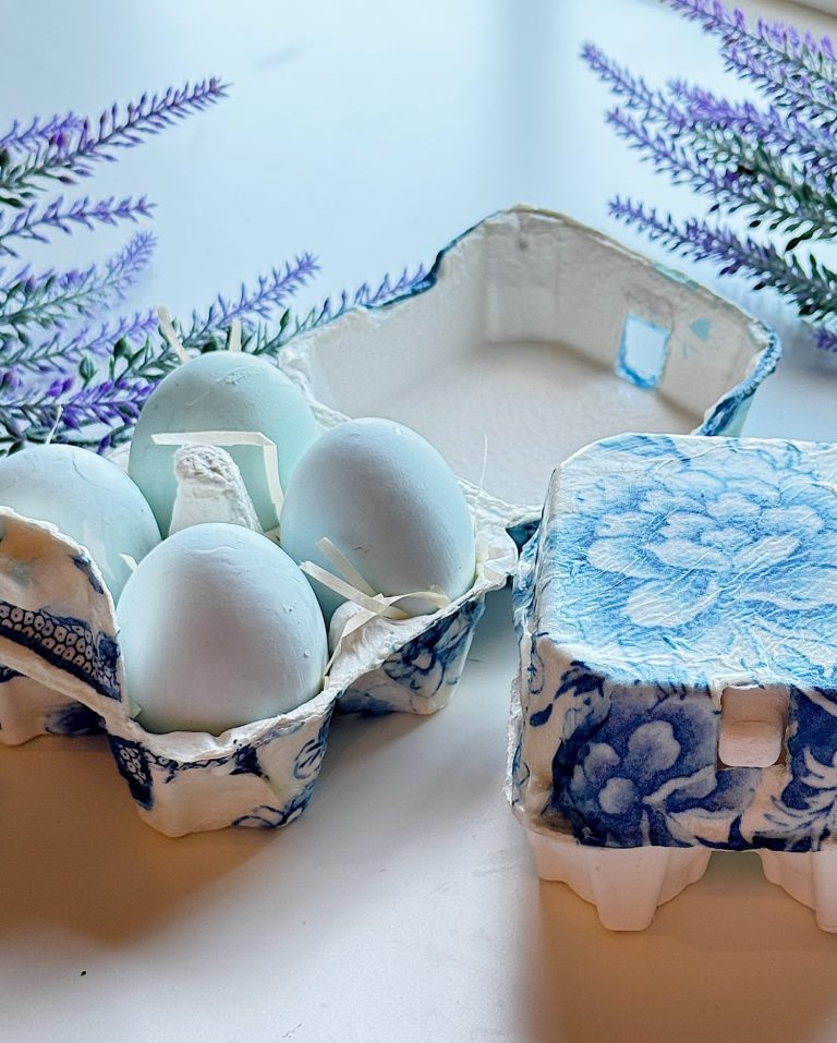 A carton of pale blue eggs in a decorative blue and white carton sits open on a white surface, with purple flowers nearby.