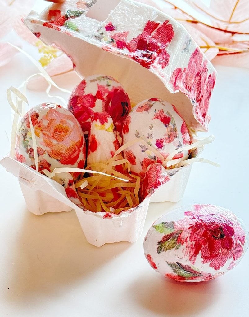 Four eggs decorated with floral patterns are placed in a white egg carton lined with straw, with one decorated egg resting outside the carton.