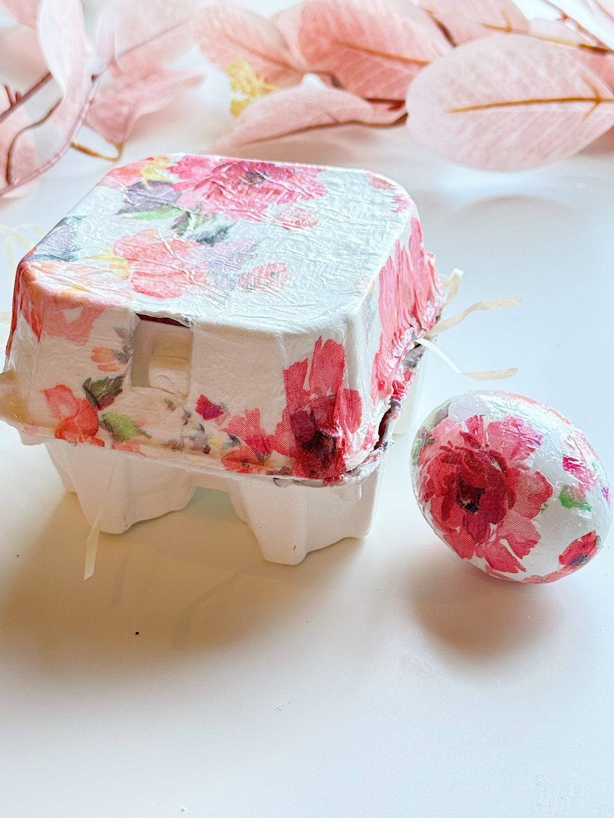 A white egg carton and an egg, both decorated with floral-patterned paper, are displayed on a white surface with pink leaves in the background.