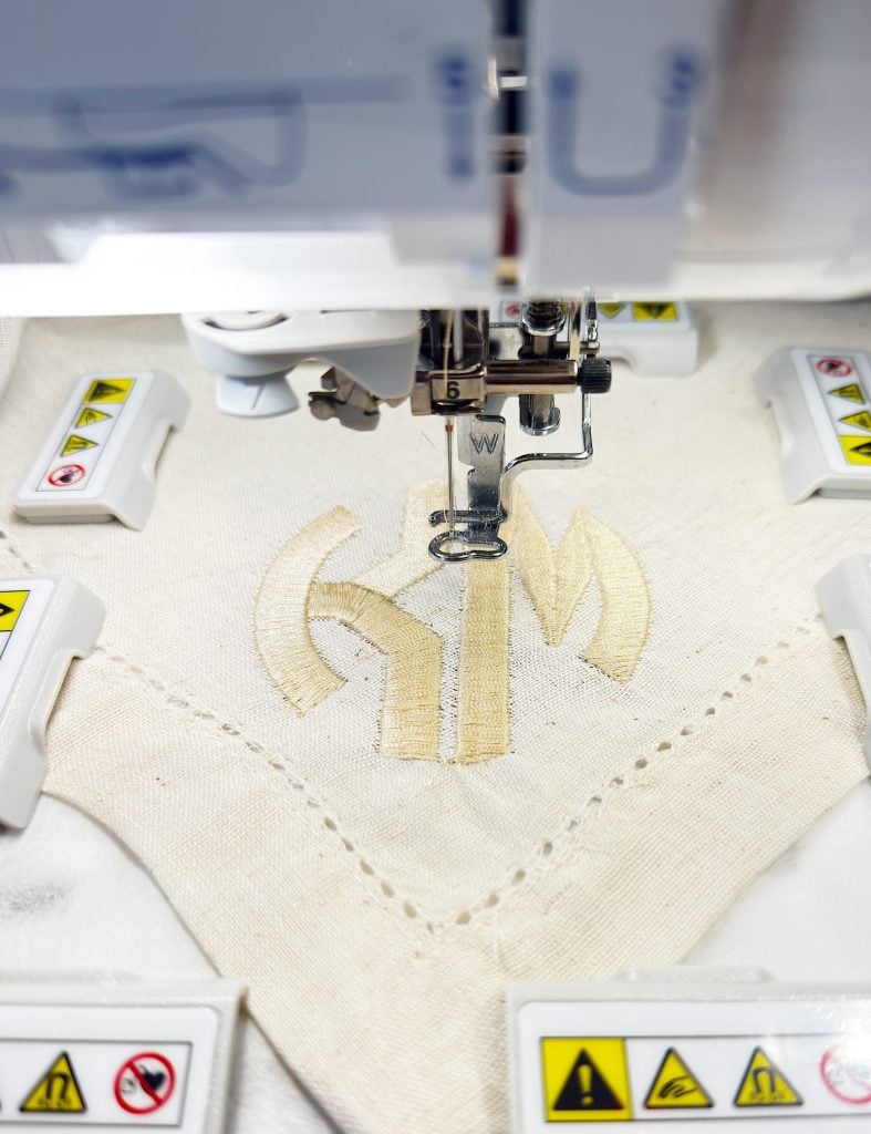 Close-up of an embroidery machine stitching a beige geometric design onto a piece of fabric, surrounded by warning labels on the machine parts.