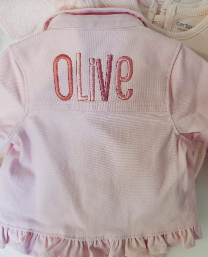 A light pink baby jacket with the name "Olive" embroidered in pink and red shades on the back.