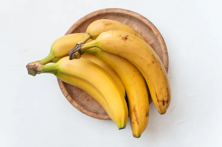 A bunch of ripe yellow bananas rests on a round wooden plate against a white background.