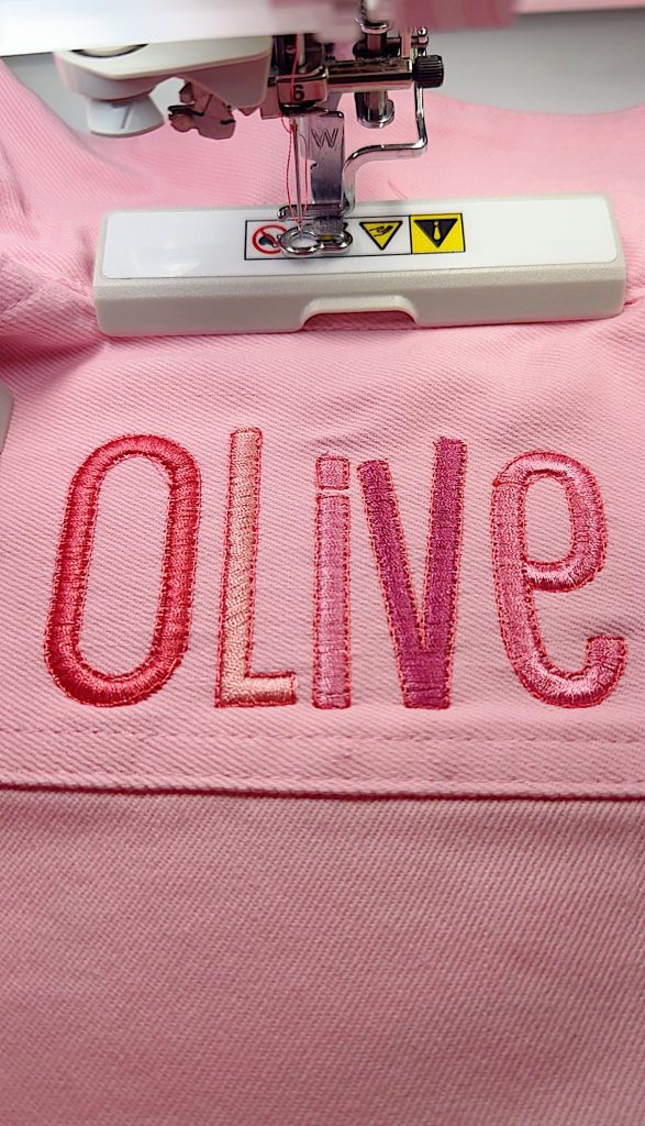 A sewing machine embroidering the word "Olive" in pink and red thread on pink fabric.