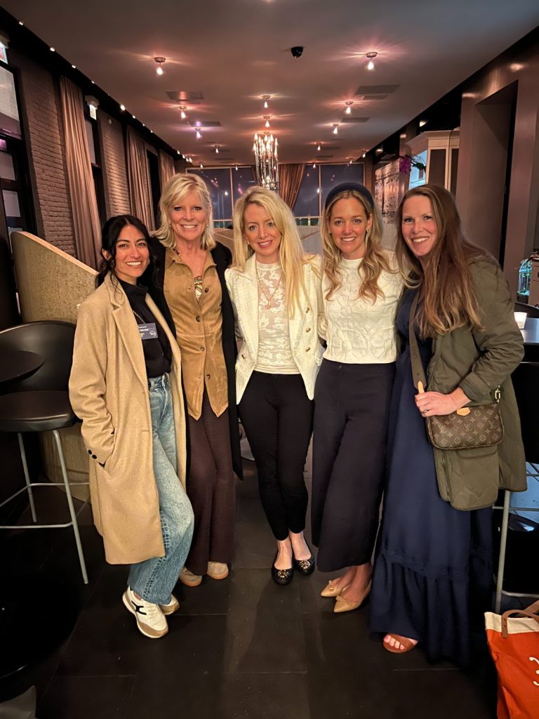 Five women stand together indoors, smiling at the camera in a modern, warmly lit lounge with dark floors and stylish decor, reflecting an inspired home atmosphere.