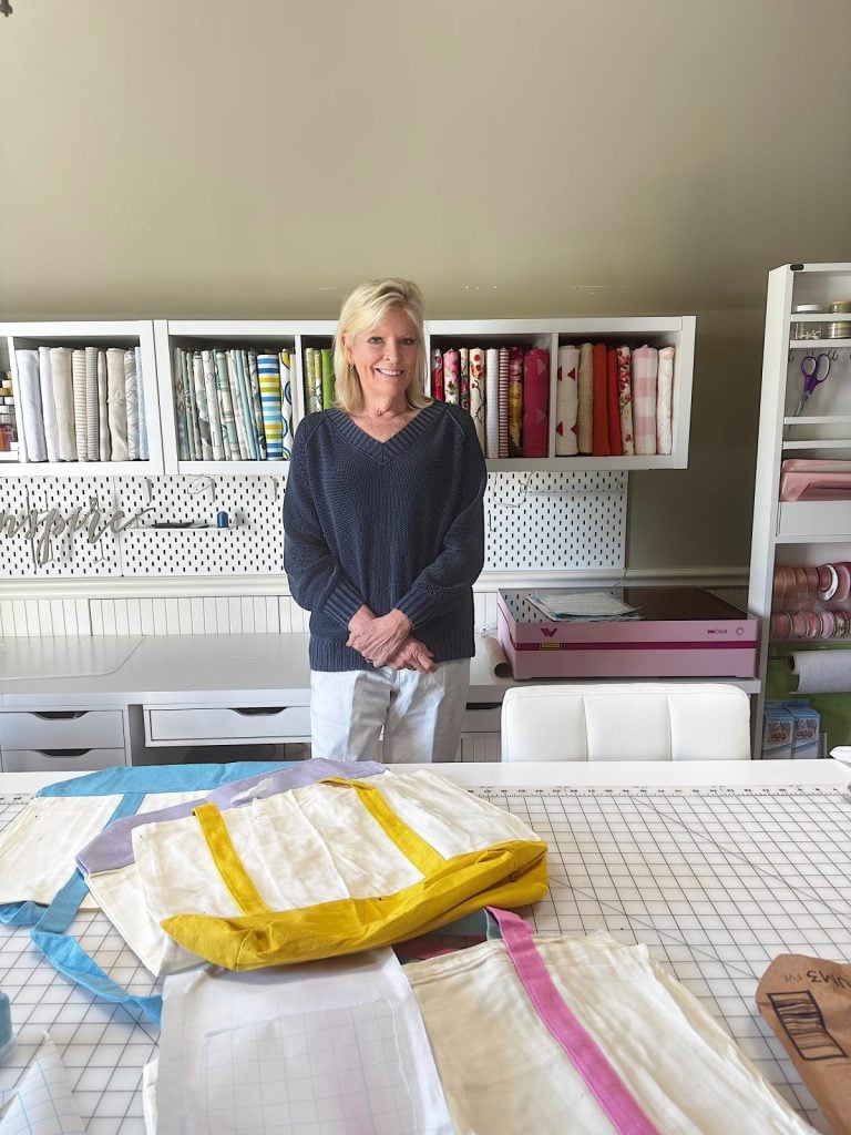 A woman stands in a sewing room behind a table with fabric tote bags, with shelves of fabric and sewing supplies in the background.