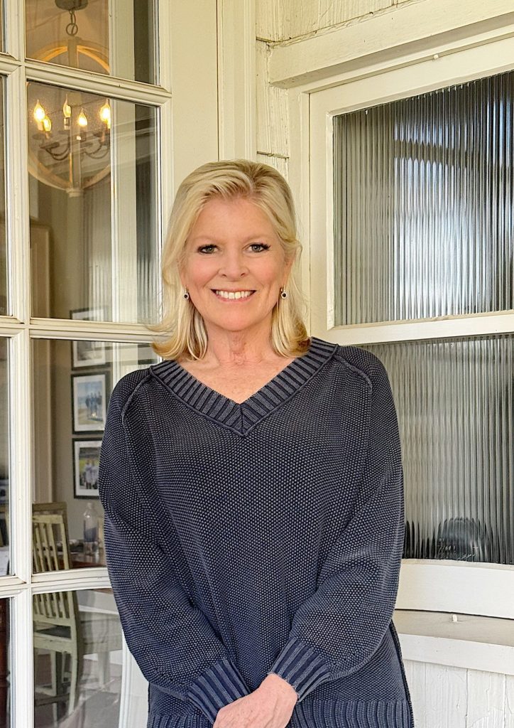 A woman with blonde hair wearing a dark sweater stands in front of a glass-paneled door, smiling at the camera.