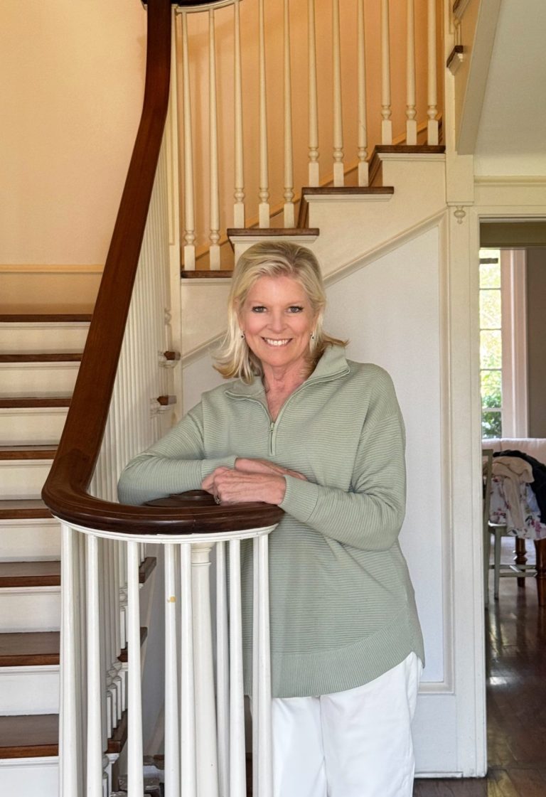 A woman in a light green sweater and white pants stands and smiles by a wooden staircase inside a house.