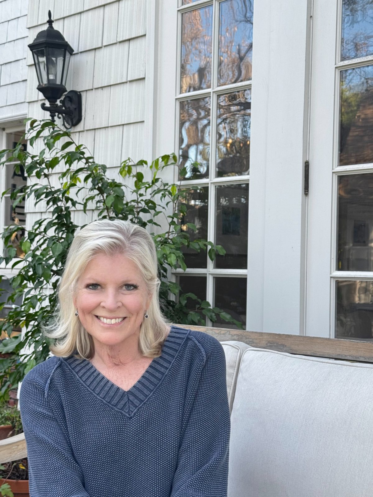 A woman with blonde hair in a blue sweater sits on an outdoor cushioned bench in front of glass doors and a leafy plant.
