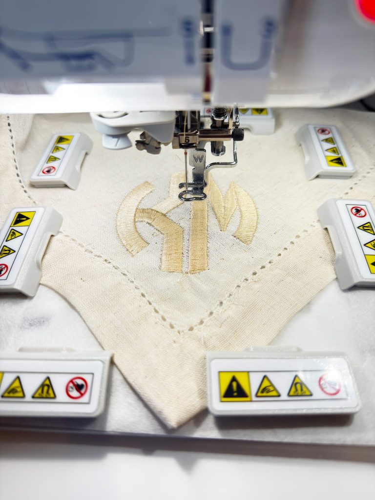 Close-up of an embroidery machine stitching a beige design onto a piece of fabric, with safety warning labels visible around the work area.