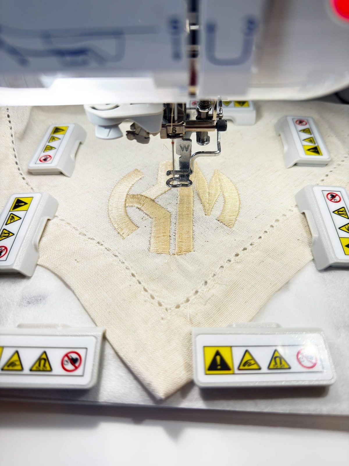 Close-up of an embroidery machine stitching a beige design onto a piece of fabric, with safety warning labels visible around the work area.