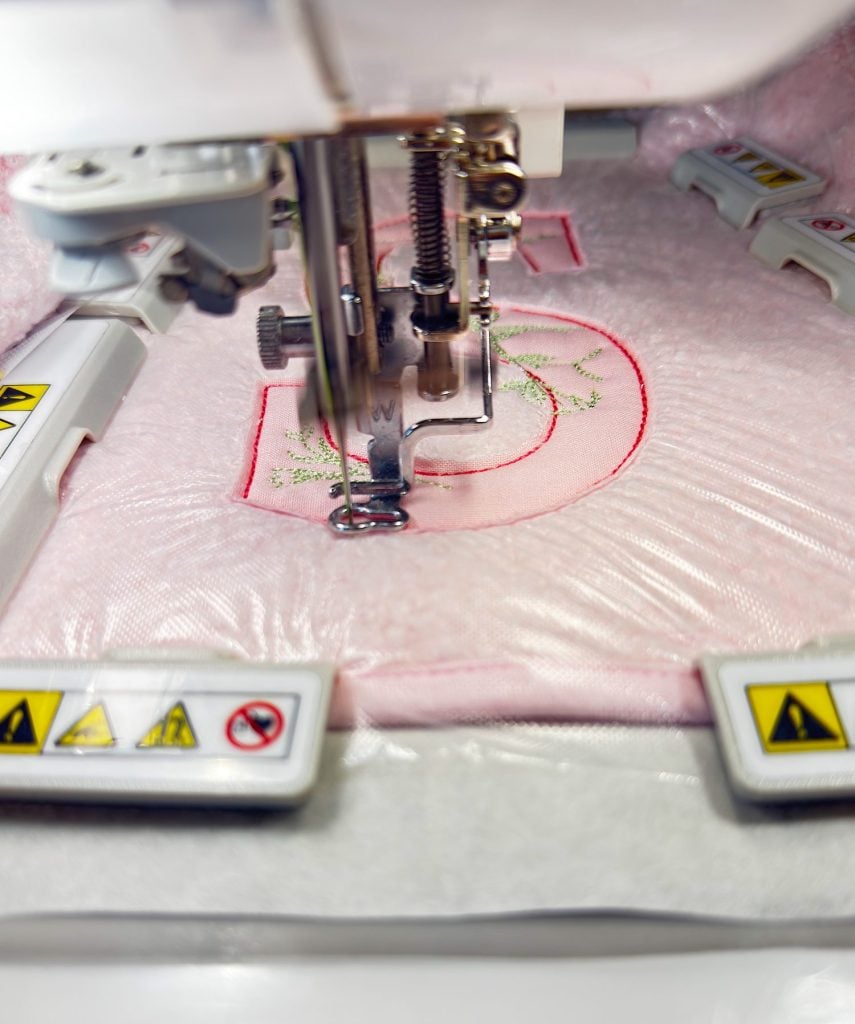 A close-up of an embroidery machine stitching a pink letter "D" with green detailing onto fabric, with safety warning labels visible on the machine.