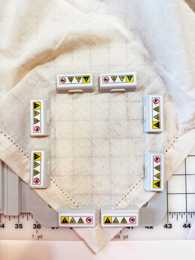Eight white rectangular blocks with caution and prohibition symbols are arranged in a square on a fabric, which is placed on a gridded cutting mat.