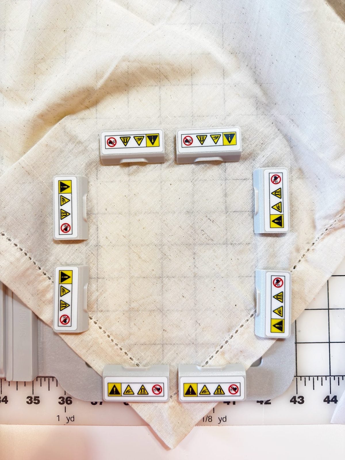 Eight white rectangular blocks with caution and prohibition symbols are arranged in a square on a fabric, which is placed on a gridded cutting mat.
