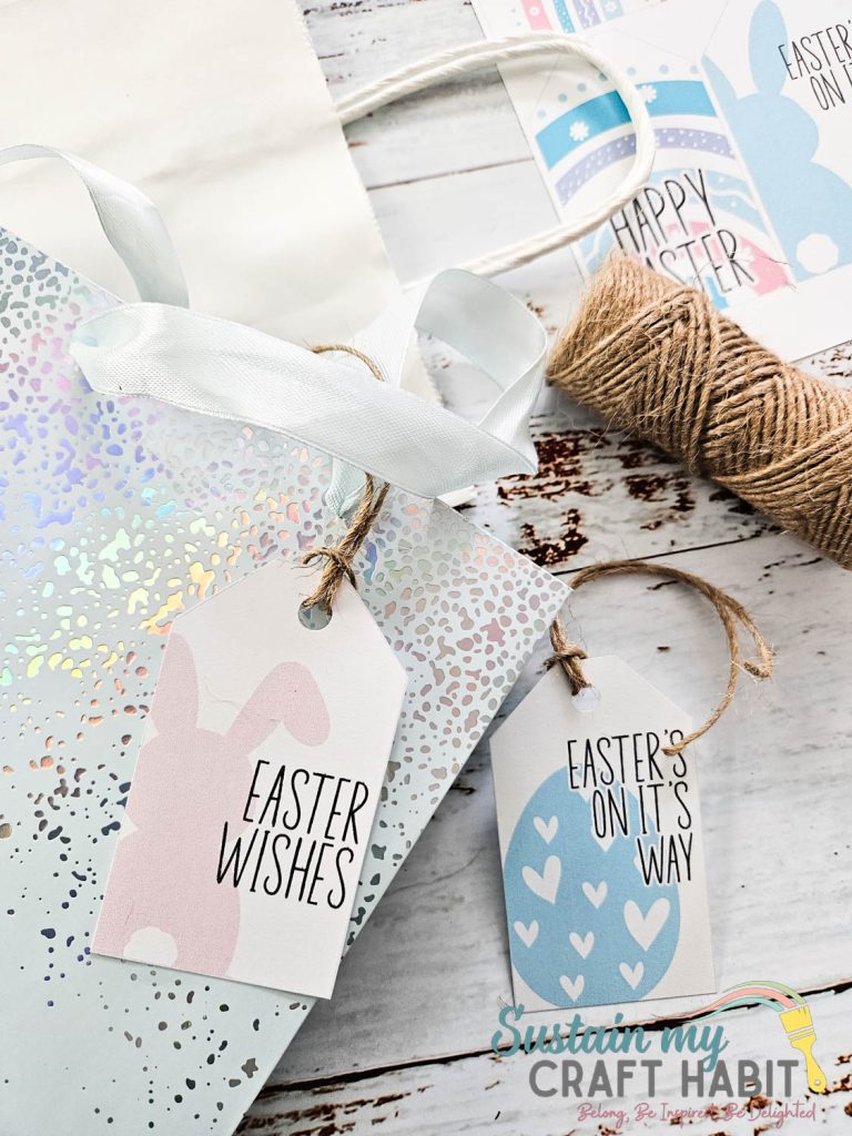 Gift bags with Easter-themed tags reading “Easter Wishes” and “Easter’s On Its Way,” alongside white ribbon and a spool of twine on a wooden surface.