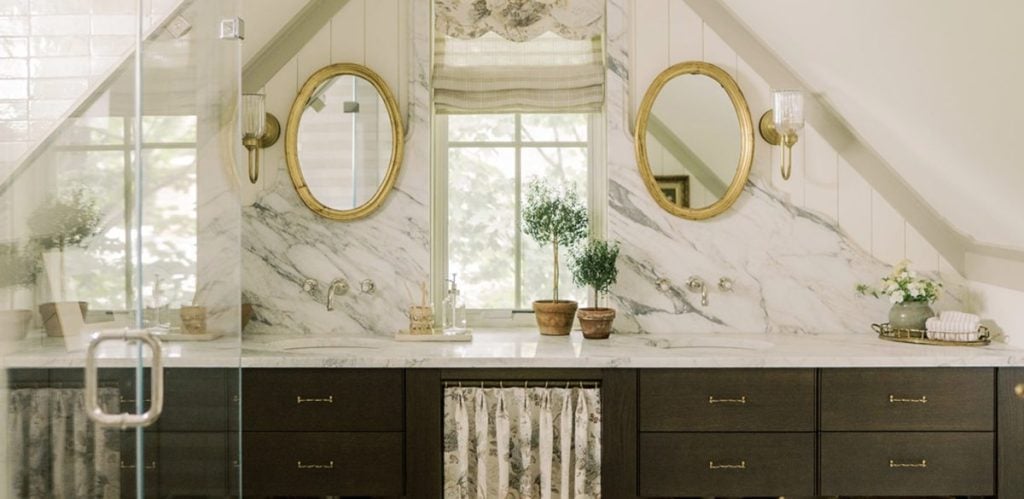 Double vanity bathroom with marble countertops, two round mirrors with gold frames, potted plants, and a window between two sloped walls.