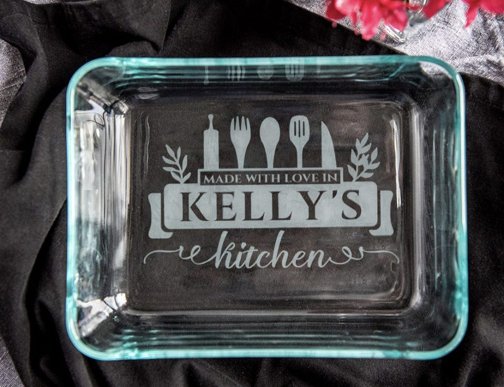 A glass baking dish with the words "Made with love in Kelly's kitchen" etched on the bottom, surrounded by utensils and decorative leaves.