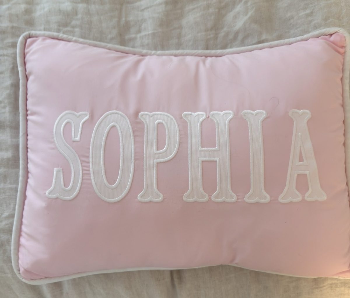 A rectangular pink pillow with white trim features the name "SOPHIA" embroidered in large white letters.