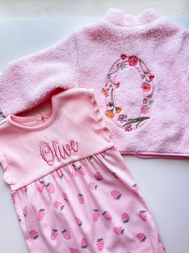 Pink baby dress with "Olive" embroidered on the chest and strawberry print, next to a pink jacket with a large floral "O" embroidered on the back.