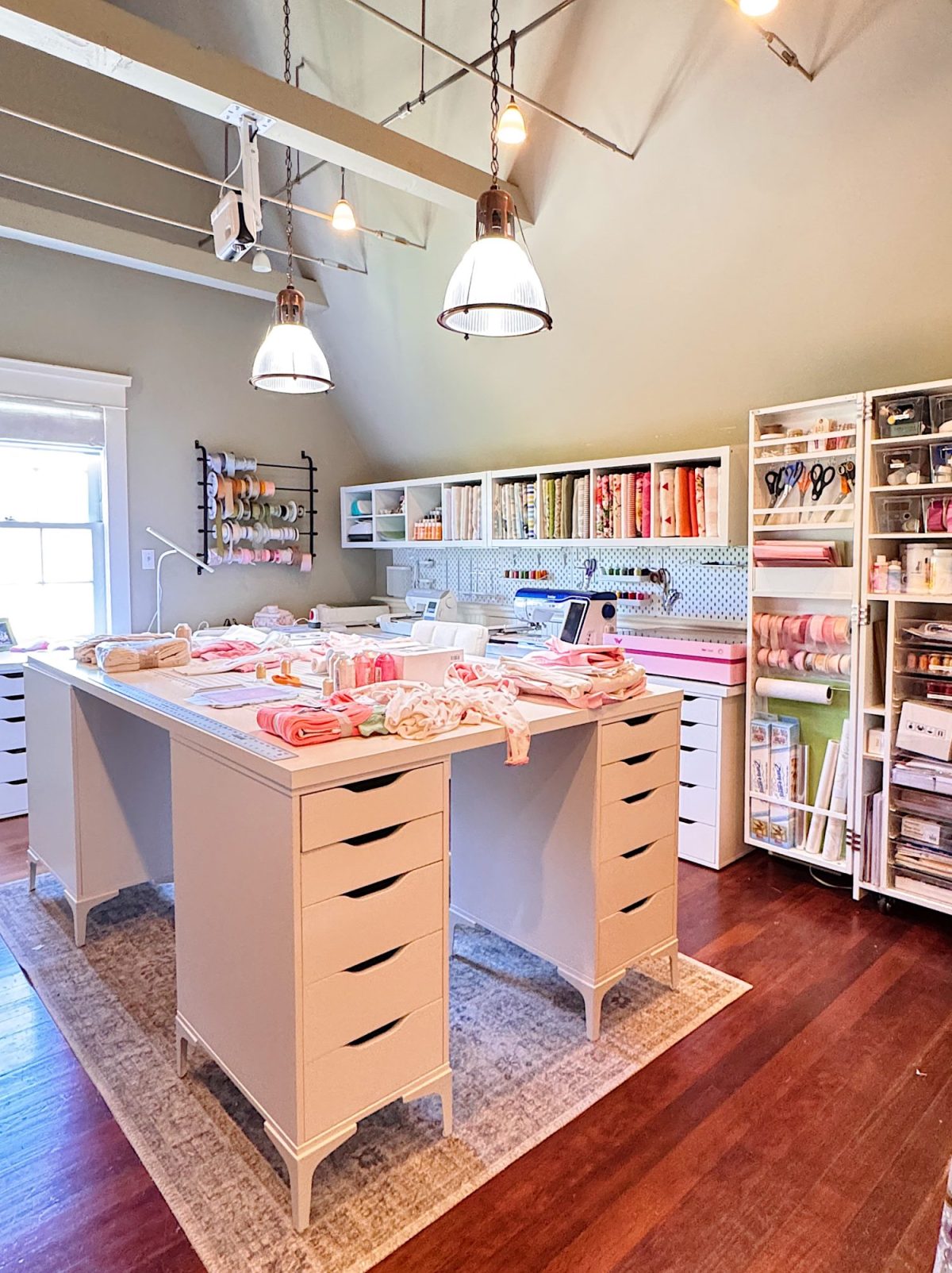 Well-lit craft room with large worktable, organized shelves of fabric, crafting supplies, and tools, featuring pink and white accents and hardwood floors.