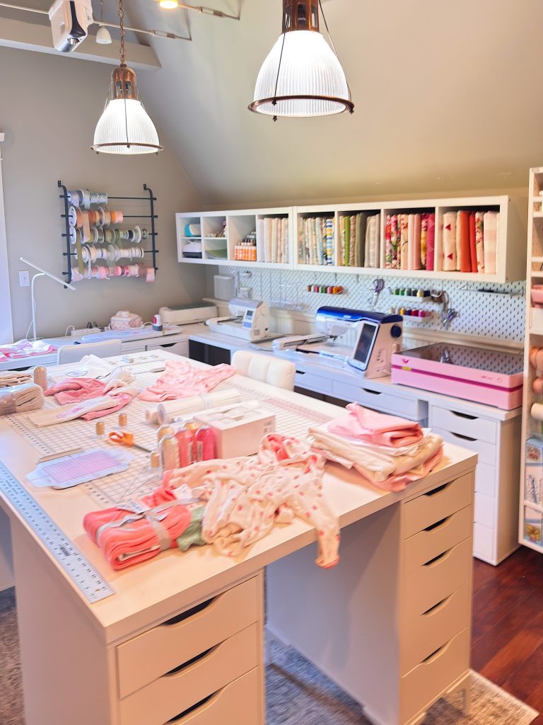 A sewing and craft room with organized fabric, ribbon spools, sewing machines, and folded materials on white worktables under pendant lights.